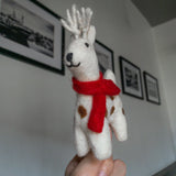 A hand holds a white wool felt Christmas reindeer ornament, presented against a gray wall with framed black and white pictures. The cheerful reindeer felt ornament has a rustic feel, featuring small brown spots, a bright red felt scarf, and white wool antlers. It also has a black stitched nose and mouth.
