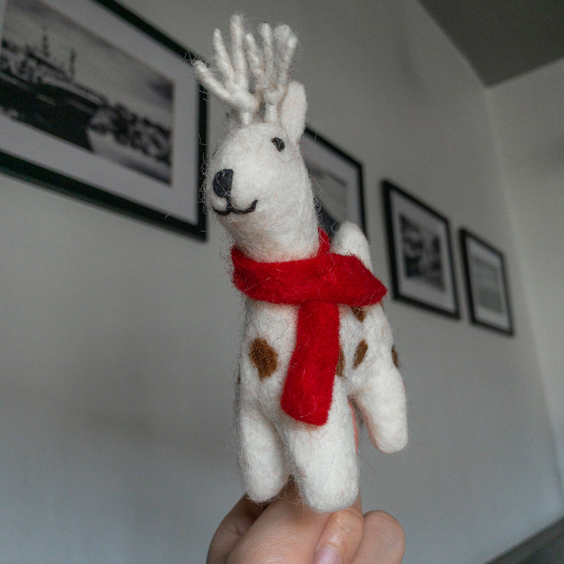 A hand holds a white wool felt Christmas reindeer ornament, presented against a gray wall with framed black and white pictures. The cheerful reindeer felt ornament has a rustic feel, featuring small brown spots, a bright red felt scarf, and white wool antlers. It also has a black stitched nose and mouth.