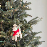 A charming needle felted reindeer Christmas ornament hangs on a festive green Christmas tree adorned with string lights. This handcrafted felt ornament features an off-white reindeer with brown spots, prominent antlers, black eyes, and a nose, completed by a vibrant red scarf. It hangs gracefully from a white cotton string.