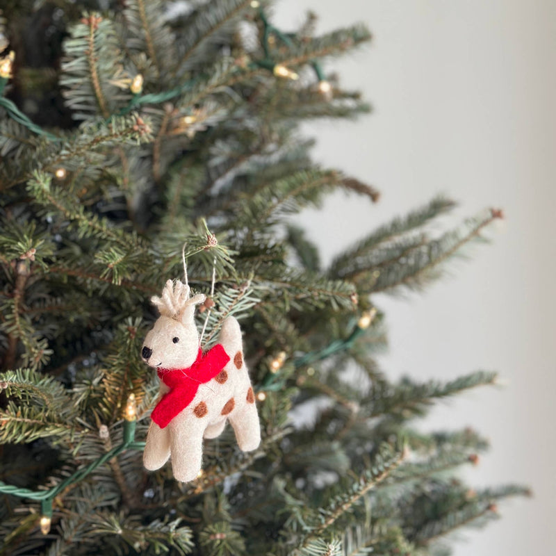 A charming needle felted reindeer Christmas ornament hangs on a festive green Christmas tree adorned with string lights. This handcrafted felt ornament features an off-white reindeer with brown spots, prominent antlers, black eyes, and a nose, completed by a vibrant red scarf. It hangs gracefully from a white cotton string.