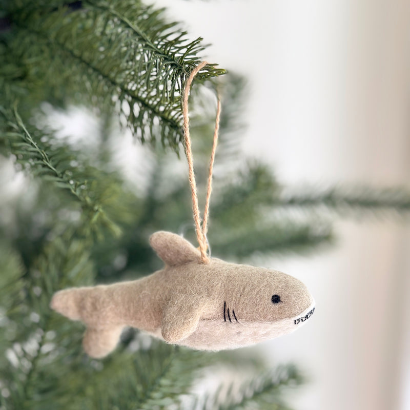 A playful needle felted shark ornament hangs from a green Christmas tree branch, set against a blurred white background. This unique Christmas tree ornament is light brown with a darker upper body and a lighter lower section. It features a dorsal fin, two side fins, a tail fin, a black eye, and black stitched details for the mouth. A brown jute string is attached, ready for display.