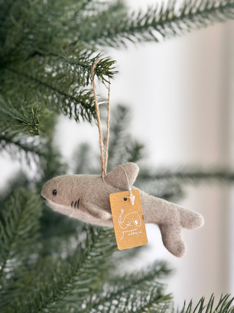 A light gray felt shark ornament hangs gracefully from the green needles of a Christmas tree. This rounded Christmas shark ornament features tiny black eyes, stitched gills, and a jute string for hanging. A small tag is also attached to this unique felt ornament, adding a festive touch.