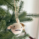A light beige felt shark ornament, with eyes, a stitched mouth showing teeth, and side fins, hangs playfully on a lush green Christmas tree. Suspended by a jute string, this unique Christmas ornament adds a cheerful and festive touch to holiday decor.