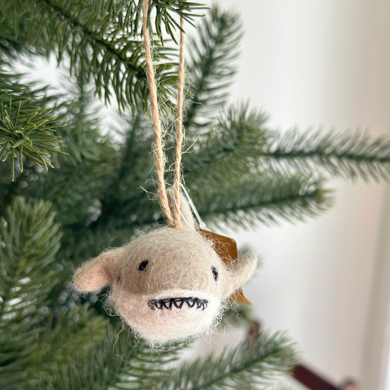 A light beige felt shark ornament, with eyes, a stitched mouth showing teeth, and side fins, hangs playfully on a lush green Christmas tree. Suspended by a jute string, this unique Christmas ornament adds a cheerful and festive touch to holiday decor.