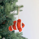 A vibrant handmade wool felt clownfish ornament hangs from a Christmas tree, evoking a playful ocean theme. The orange clownfish ornament features distinct white stripes and small black eyes, crafted from soft sheep wool. It is suspended by a rustic jute string, adding a whimsical touch to the holiday decor.