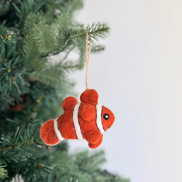 A vibrant handmade wool felt clownfish ornament hangs from a Christmas tree, evoking a playful ocean theme. The orange clownfish ornament features distinct white stripes and small black eyes, crafted from soft sheep wool. It is suspended by a rustic jute string, adding a whimsical touch to the holiday decor.