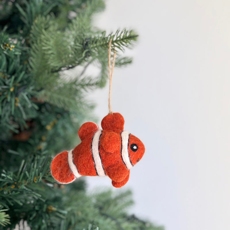 A vibrant handmade wool felt clownfish ornament hangs from a Christmas tree, evoking a playful ocean theme. The orange clownfish ornament features distinct white stripes and small black eyes, crafted from soft sheep wool. It is suspended by a rustic jute string, adding a whimsical touch to the holiday decor.