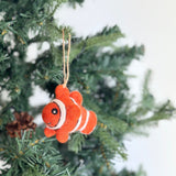 A vibrant wool felt clownfish ornament hangs playfully on a Christmas tree, suspended by a jute string. This whimsical clownfish features a primary orange body with distinct white stripes and a small, round black eye. It's nestled among the green pine needles, with a pine cone visible nearby, adding a rustic touch to the holiday display.