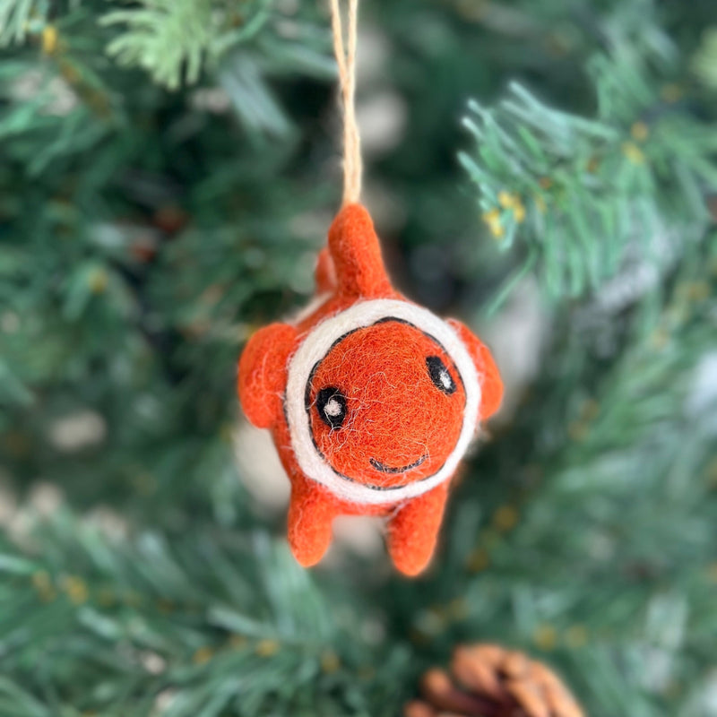 A cheerful, handmade wool felt clownfish ornament hangs from a blurred green Christmas tree. This vibrant orange clownfish features distinctive white stripes and black accents around its eyes and mouth. Crafted from soft sheep wool felt, the unique Christmas ornament includes a jute string for hanging.