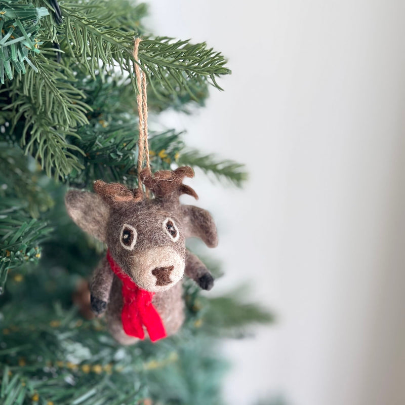 A cute Christmas Reindeer felt ornament hangs from a festive Christmas tree. This charming felted reindeer ornament features a light gray body, a dark brown nose, small antlers, and a bright red scarf, suspended by a jute string amidst the green needles and string lights.