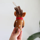 A handcrafted felt reindeer ornament is delicately held by a female hand against a soft white background. This charming Christmas tree ornament features a light brown body with white spots, dark brown felt antlers, and a red cotton string around its neck, ready for festive display.