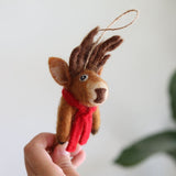 A handcrafted felt reindeer ornament is held closely by a female hand against a white background. This cute Christmas reindeer features fluffy brown antlers, a snugly wrapped red scarf, perky ears, and a charming expression, all crafted from soft, wool-like felt. A jute string loop is attached at the top for hanging.
