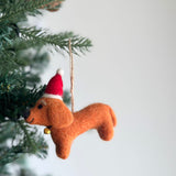 A brown felt dachshund ornament with a playful white felt bone center hangs from a green Christmas tree. This detailed, handmade felt ornament features the dachshund's distinctive long body and short legs, set on a white felt frame. A jute string allows for easy hanging, adding a whimsical touch to any festive display.