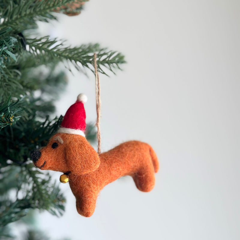 A brown felt dachshund ornament with a playful white felt bone center hangs from a green Christmas tree. This detailed, handmade felt ornament features the dachshund's distinctive long body and short legs, set on a white felt frame. A jute string allows for easy hanging, adding a whimsical touch to any festive display.