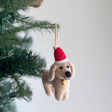 A festive handmade felt dachshund ornament hangs gracefully from a Christmas tree branch. This unique Christmas ornament features four adorable felt dachshunds in cream, khaki, brown, and black, arranged on a white felt frame. A playful felt bone is centered on the frame, adding to its joyful appeal. It hangs by a jute string.