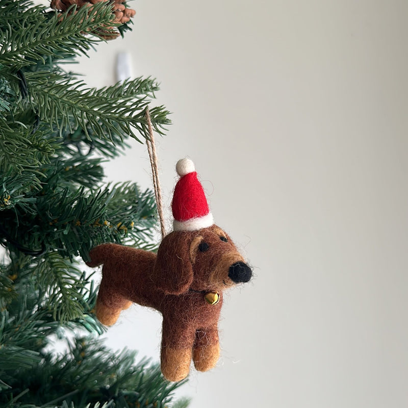 A single brown felt dachshund ornament hangs from a snow-dusted green Christmas tree branch, set against a plain white wall. This festive felt dachshund ornament features darker brown details, a black nose, and dark eyes, complete with a brown jute string for hanging.