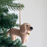 A light-brown felt dachshund ornament hangs playfully from a Christmas tree branch. This handcrafted felt dachshund ornament features a soft, textured surface resembling fur, with small dark eyes and a black, wooly nose. Tiny legs, a tail, and a small golden bell tied with a black string around its neck add to its whimsical charm. It hangs from a rustic jute string against a clean white background.