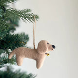 Felt Dachshund Christmas tree ornaments are charmingly suspended from a green branch, set against a softly blurred holiday background. This unique felt ornament displays four adorable dachshunds in cream, khaki, brown, and black, arranged on a white felt frame. A playful felt bone accents the center.