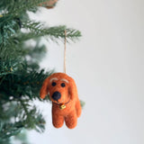 A felt dachshund ornament hangs playfully from a green Christmas tree branch. The charming dachshund features small dark eyes, a black nose, and floppy ears, with a tiny golden bell around its neck. A jute string loop allows for easy hanging, completing this delightful Christmas tree ornament.