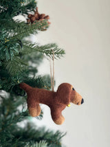 Felt dachshund ornaments are playfully displayed on a lush green Christmas tree. This charming ornament features four felt dachshunds in cream, khaki, brown, and black, surrounding a felt bone within a white felt frame. A jute string allows it to hang among the needled branches and a natural pine cone, against a soft light gray background.