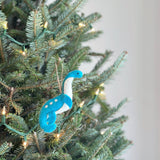 A wool-felt plesiosaur dinosaur ornament is nestled among the green branches of a Christmas tree adorned with warm white lights. This playful felt ornament features a turquoise body, a long, curving white neck, and a small head, accented with white dots. A jute string is attached for hanging this unique Christmas decoration.