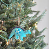 A charming hand-stitched wool-felt Stegosaurus ornament hangs from a Christmas tree adorned with warm, inviting lights. This delightful wool-felt dinosaur features a light blue body, accented by dark gray scales and evenly spaced white and dark blue dots. A rustic jute string enhances its handmade appeal as it dangles from the branch.