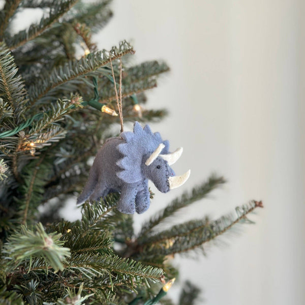 A light gray felt dinosaur triceratops ornament hangs on a festive green Christmas tree adorned with tiny light bulbs. This hand-stitched wool felt triceratops features a light gray body, a textured frill, and white horns, showcasing detailed craftsmanship. A jute string is attached to the top for easy hanging.