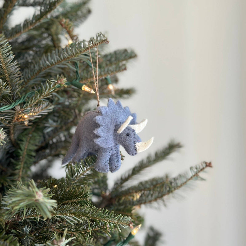 A light gray felt dinosaur triceratops ornament hangs on a festive green Christmas tree adorned with tiny light bulbs. This hand-stitched wool felt triceratops features a light gray body, a textured frill, and white horns, showcasing detailed craftsmanship. A jute string is attached to the top for easy hanging.