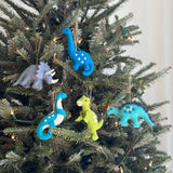 A grey wool-felt Triceratops dinosaur Christmas ornament hangs prominently on a well-lit Christmas tree, surrounded by other unique wool-felt dinosaur ornaments. Crafted from 100% New Zealand sheep wool, this charming triceratops features four legs, three horns, and a tail, adding a playful, prehistoric touch to the holiday decor.