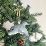 A charming needle felted dolphin ornament hangs on a Christmas tree, visible with green needles and a partial pine cone. This unique Christmas tree ornament is light blue, fading to gray at its tail, detailed with a small black eye, and suspended by a rustic jute string.