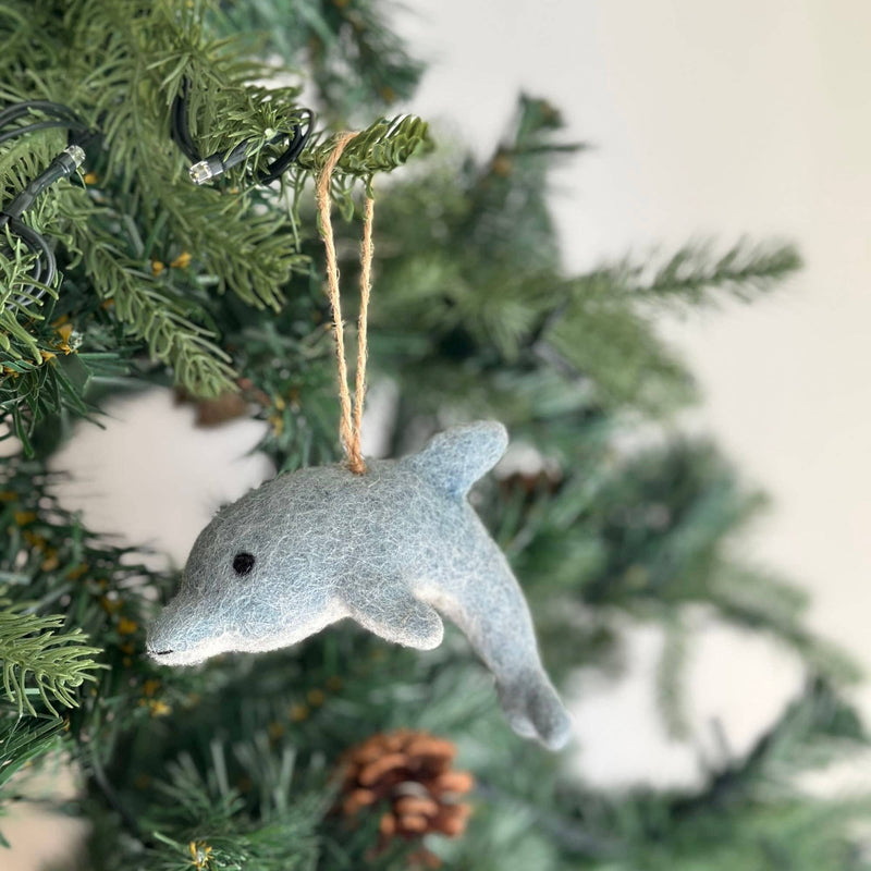 A charming needle felted dolphin ornament hangs on a Christmas tree, visible with green needles and a partial pine cone. This unique Christmas tree ornament is light blue, fading to gray at its tail, detailed with a small black eye, and suspended by a rustic jute string.