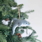 A light gray felt dolphin Christmas ornament hangs gracefully from a vibrant green Christmas tree branch, adding a calm and festive charm. The handcrafted felt ornament features a streamlined body with a small dorsal fin, a pointed snout, and a tiny black eye, creating a smooth and elegant design.