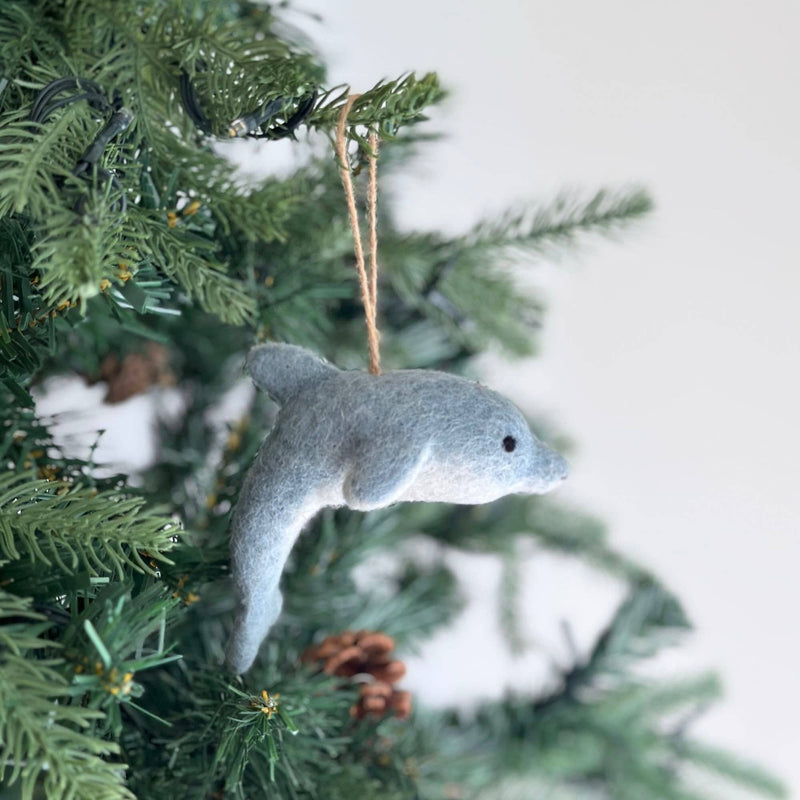 A light gray felt dolphin Christmas ornament hangs gracefully from a vibrant green Christmas tree branch, adding a calm and festive charm. The handcrafted felt ornament features a streamlined body with a small dorsal fin, a pointed snout, and a tiny black eye, creating a smooth and elegant design.