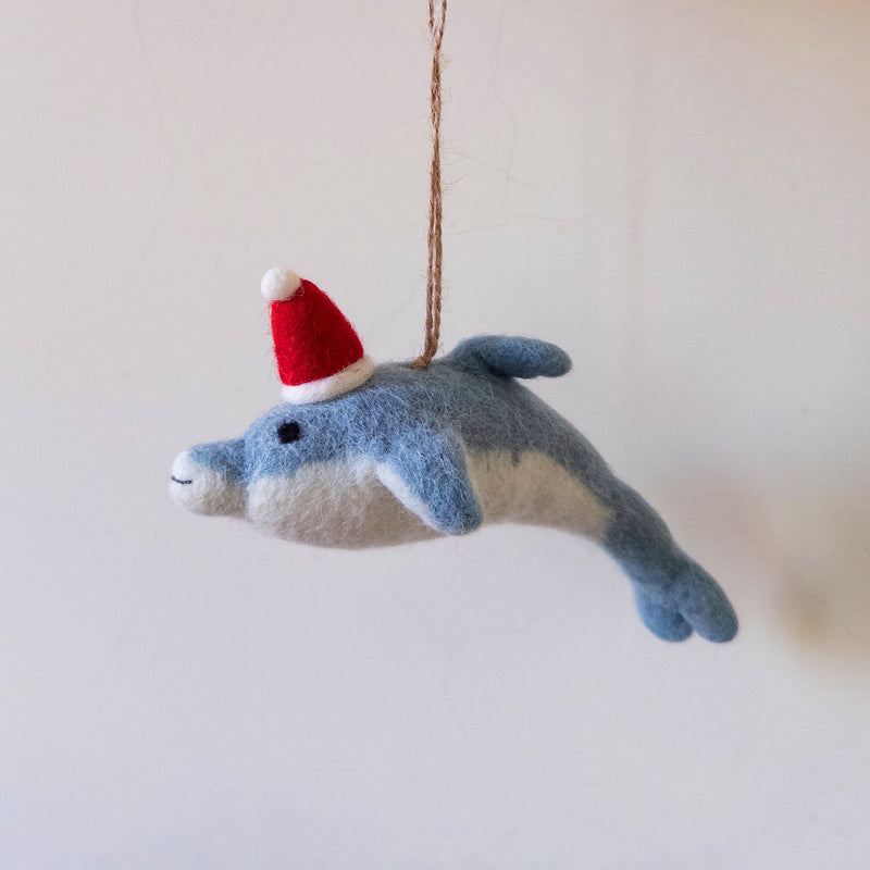 A whimsical felt dolphin ornament, ready for the Christmas tree, is presented against a plain white background. This cheerful handmade felt ornament is light blue with a white belly, featuring small black eyes, a stitched mouth, and a jaunty red and white Santa hat. A jute string is attached for easy hanging.