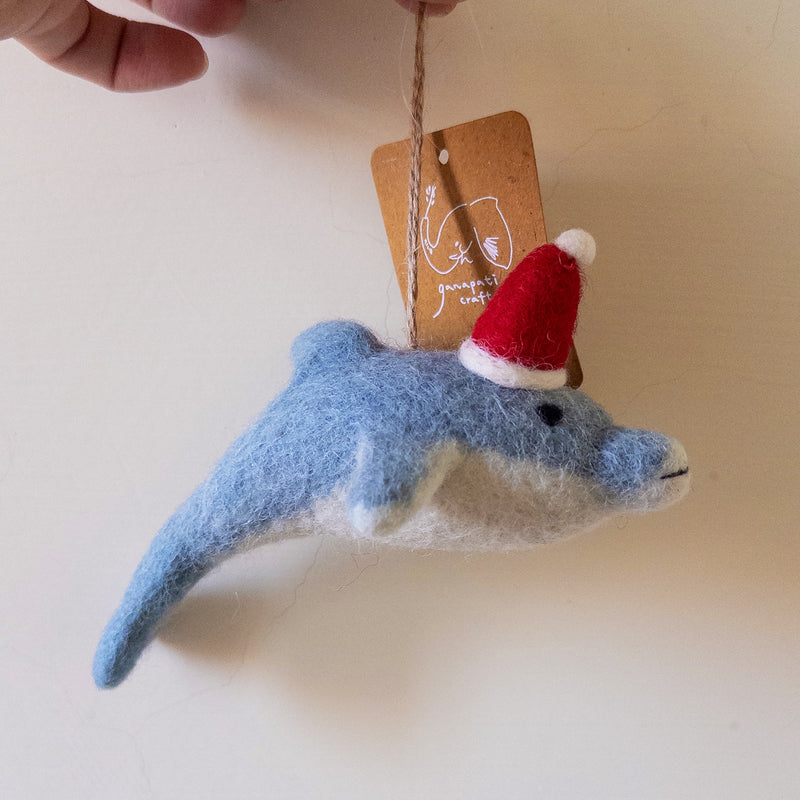 A handcrafted felt dolphin ornament is gently held by a hand against a muted background. The light blue dolphin features a white underbelly, a black eye, and a stitched mouth, crafted from soft wool felt. A festive red Santa hat with a fluffy white pompom sits jauntily on its head, and it hangs from a jute string attached to a light brown product tag.