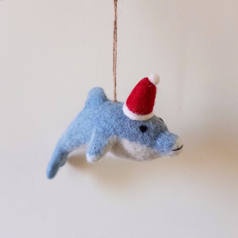 A needle felted dolphin ornament hangs by a jute string against a pale background. This light blue dolphin features a white underbelly and a charming red and white Santa hat. Its small black dot eye and soft felt texture highlight its handcrafted appeal, creating a whimsical, festive mood.