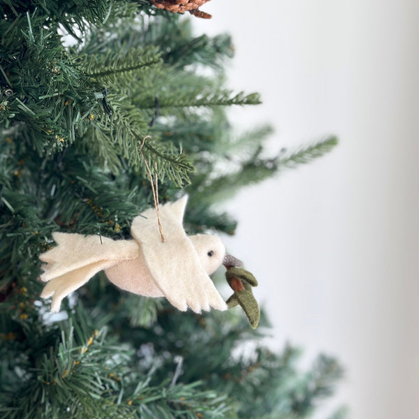 A pure white wool felt dove ornament, holding a delicate green olive twig in its beak, hangs on a lush green Christmas tree. This handcrafted felt ornament features detailed wings and a small dark eye, evoking a peaceful and festive holiday atmosphere. A jute string is attached for hanging.