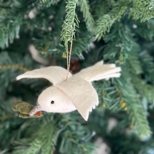 A white wool felt dove ornament, holding a green olive twig in its beak, is suspended on a brightly lit Christmas tree. The Christmas tree features soft green needles and subtle warm lights. A jute string is attached to the top of the dove for hanging.
