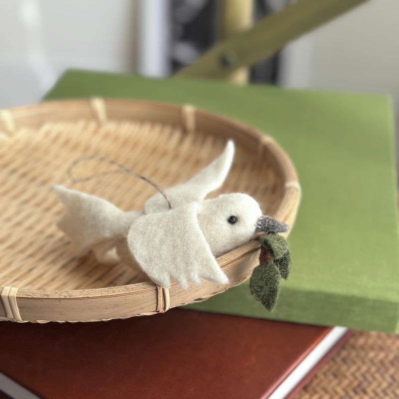 A pure white wool felt dove ornament with an olive twig rests peacefully in a round woven basket, which is placed atop a dark red book and a green box. This charming felt dove ornament features fluffy wings and tiny black eyes, holding a small green felt olive twig in its beak. A jute string is attached, ready for hanging.