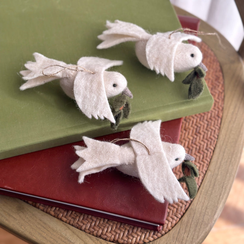 Three handcrafted wool felt dove ornaments, each with a delicate olive twig in its beak, are artfully arranged on a stack of red and green books. The pure white felted doves feature a jute string for hanging and are displayed on a round, woven surface, creating a tranquil and serene atmosphere.