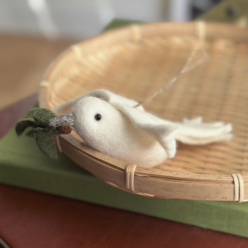 A pure white wool felt dove ornament with an olive twig in its beak is nestled inside a round, light brown woven basket. The delicate dove features a small black eye and green leaves on the twig, with a silver string attached for hanging. The basket rests on muted green and reddish-brown surfaces, creating a serene display.