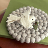 A pristine white handmade wool felt dove ornament, holding an olive twig, gracefully rests on a circular pad made of varying gray wool felt balls. The dove features carefully shaped wings, and a small olive twig in its beak symbolizes peace. A jute string is attached, ready for hanging. Its soft texture evokes a calm, handcrafted aesthetic.