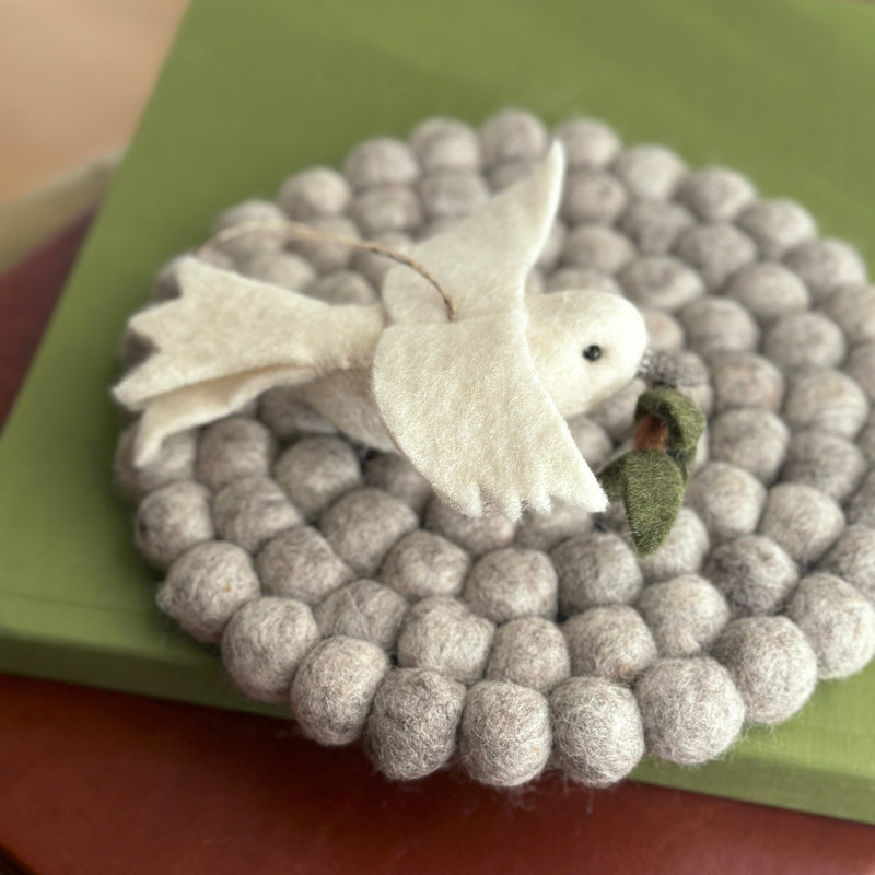 A pristine white handmade wool felt dove ornament, holding an olive twig, gracefully rests on a circular pad made of varying gray wool felt balls. The dove features carefully shaped wings, and a small olive twig in its beak symbolizes peace. A jute string is attached, ready for hanging. Its soft texture evokes a calm, handcrafted aesthetic.