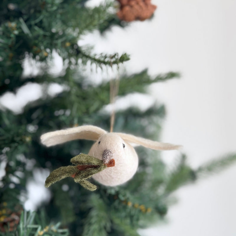 A handcrafted wool felt dove ornament with an olive twig in its beak hangs on a snow-covered Christmas tree branch, creating a serene, festive scene. The pure white dove ornament, suspended by a jute string, features a green felt olive twig. This unique Christmas ornament brings a peaceful, winter atmosphere.