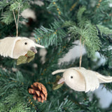 Two wool felt dove ornaments with olive twigs hang gracefully on a vibrant green Christmas tree. Each handcrafted ornament features a pure white body and wings, complemented by a small grey beak holding a green olive twig. Jute strings suspend the doves among the branches, where a brown pine cone adds a rustic, peaceful touch to the holiday display.