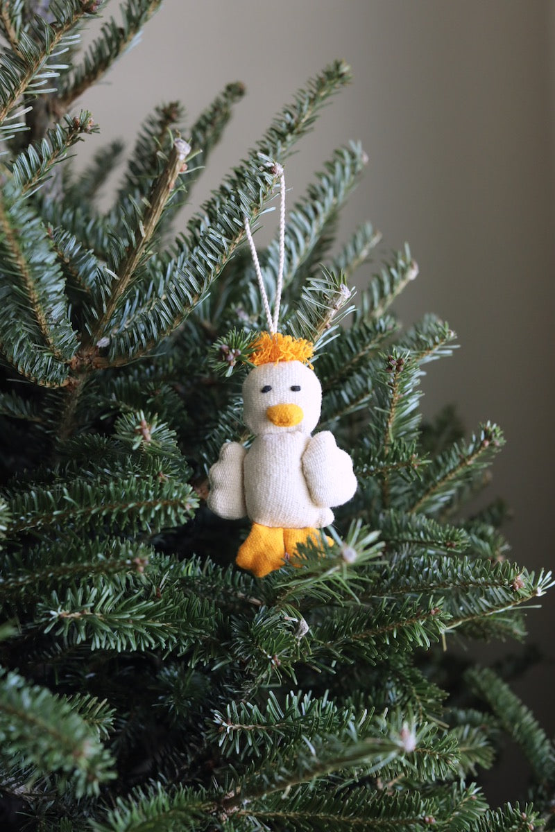 A handmade off-white felt duck ornament is nestled among the vibrant green needles of a Christmas tree. This soft duck ornament features a bright yellow beak, feet, and a tuft of yellow felt hair on its head. A white cotton string allows this endearing duck Christmas ornament to dangle gracefully from the branch, adding a whimsical touch.
