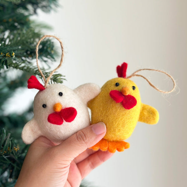 Two wool felt Easter chick ornaments are held by a female hand, displayed up close against a festive Christmas tree. One white chick ornament has a red wattle and orange beak, while the yellow chick ornament features a red wattle, beak, and orange feet. These charming, egg-shaped, needle-felted ornaments feature black wings and a sturdy hemp string, crafted from soft wool.