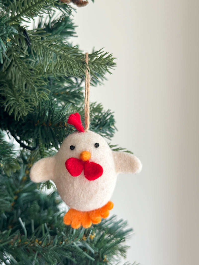 A needle felted Easter chick ornament hangs from the vibrant green branches of a Christmas tree, radiating playful holiday charm. Crafted from soft white wool felt, this cheerful chick features black eyes, an orange beak, a bright red comb, and distinctive bow-like wings. Orange felted feet peek from its bottom, with a sturdy hemp string attached for hanging.