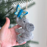 A gray felt elephant ornament with a butterfly is gently held by a hand near a green leafy branch, against a soft, neutral background. The handmade felt elephant sits with a light and dark blue butterfly, featuring yellow dots, perched whimsically on its trunk. A jute string for hanging completes this charming unique Christmas ornament.