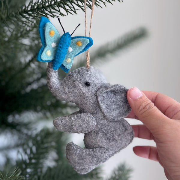 A handcrafted felt elephant ornament with a light blue butterfly is suspended by a jute string from a Christmas tree branch. The gray felt elephant features floppy ears, a curled trunk, and black eyes. A whimsical light blue butterfly with yellow and white dots rests on its trunk.